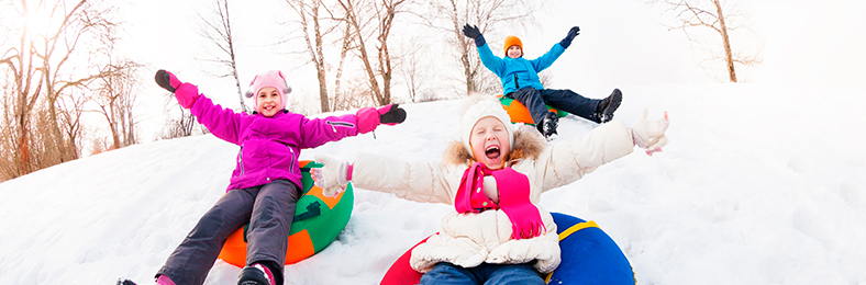 Kolme lasta talvisessa maisemassa mäenlaskussa värikkäissä vaatteissa.